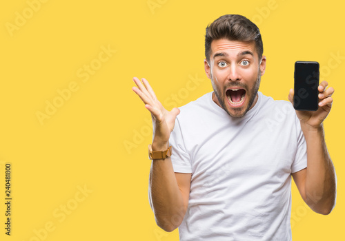 Young handsome man showing smartphone screen over isolated background very happy and excited, winner expression celebrating victory screaming with big smile and raised hands