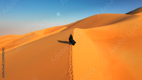 Photography Aerial view from a drone flying next to a woman in abaya (United Arab Emirates traditional dress) walking on the dunes in the desert of the Empty Quarter
