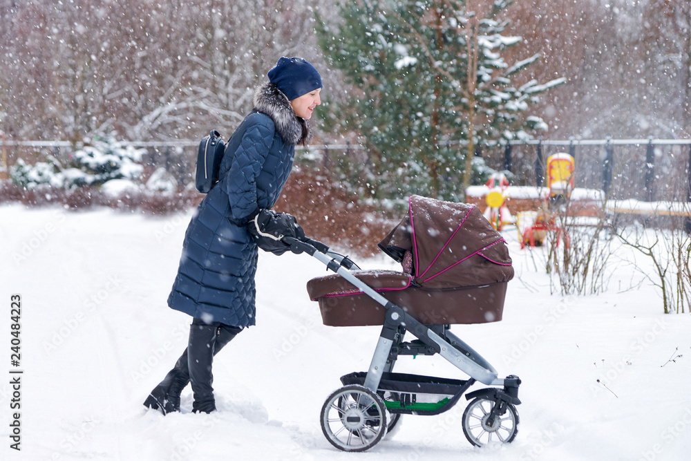 Young mother with a baby carriage in the park during snowfall