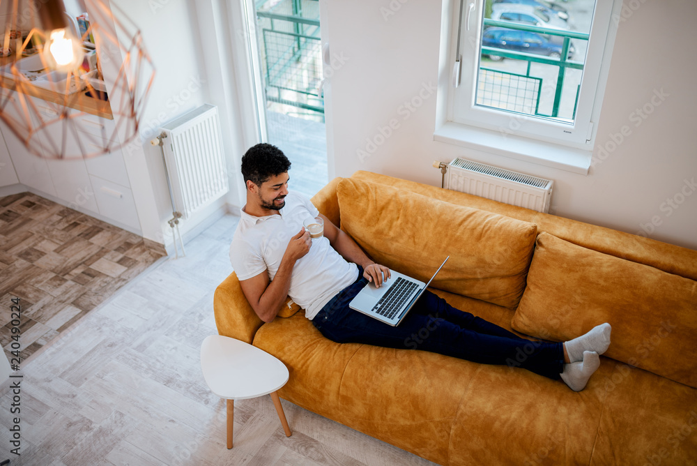 High angle image of african american man relaxing on sofa with laptop and coffee. High angle image of african american man relaxing on sofa with laptop and coffee.