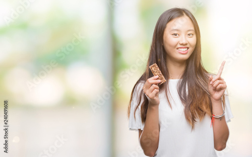 Young asian woman eating chocolate energetic bar over isolated background surprised with an idea or question pointing finger with happy face  number one