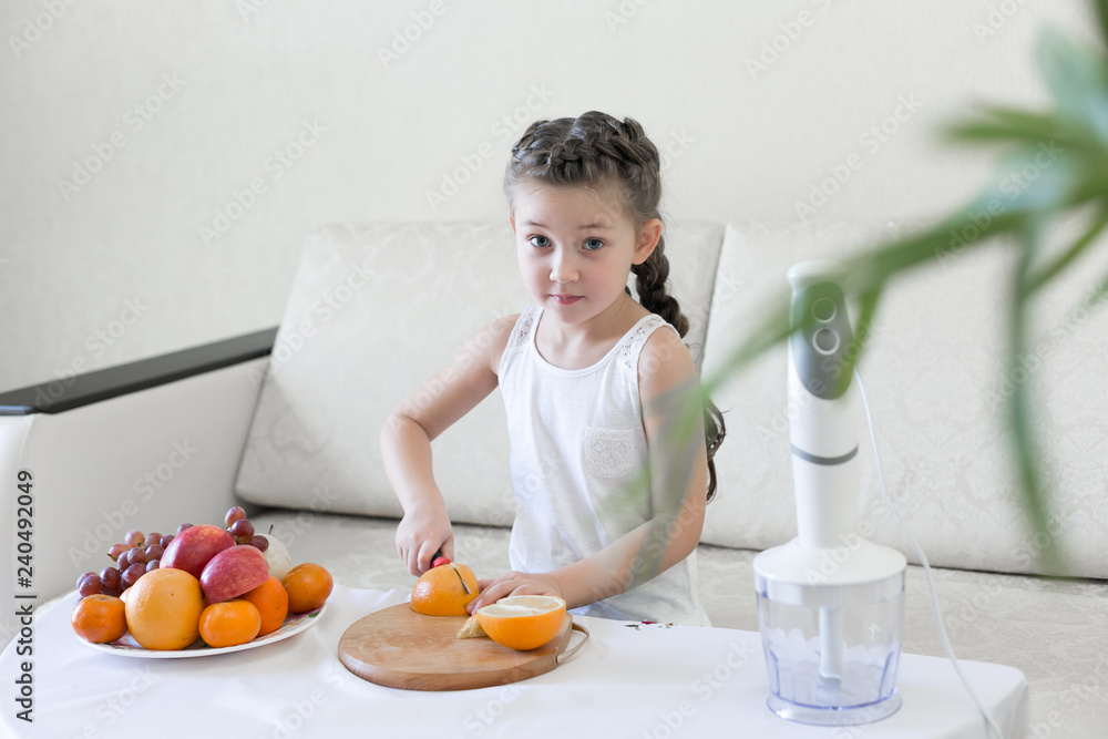 The child slices an orange with a knife. The girl cuts with a knife the fruit for making a cocktail.