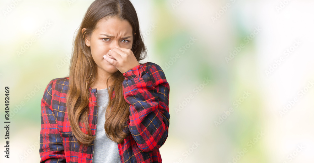 Naklejka premium Young beautiful brunette woman wearing a jacket over isolated background smelling something stinky and disgusting, intolerable smell, holding breath with fingers on nose. Bad smells concept.