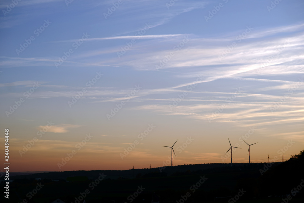 Silhouetten von Windrädern vor dem Abendhimmel / Die Silhouetten von Windrädern auf einem bewaldeten Hügel am Abend. .. Silhouetten von Windrädern vor dem Abendhimmel / Die Silhouetten von Windrädern auf einem bewaldeten Hügel am Abend. ..