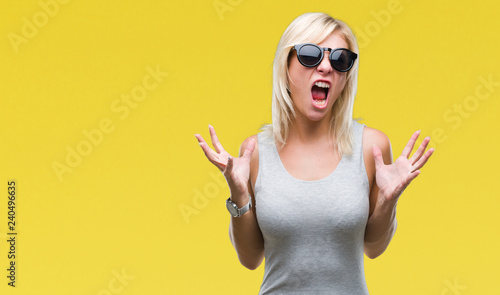 Young beautiful blonde woman wearing sunglasses over isolated background crazy and mad shouting and yelling with aggressive expression and arms raised. Frustration concept.
