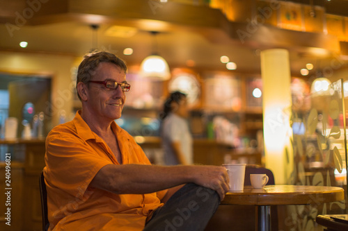 Man holding mug of coffee with a smile enjoying life in a Cafe wearing a orange shirt