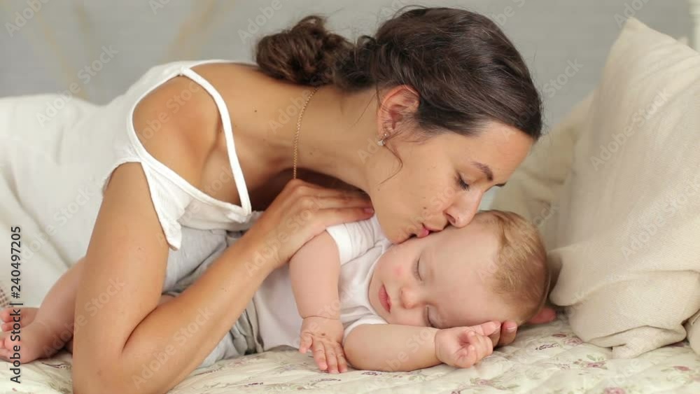 Happy mom is lying with her newborn sleeping baby in bed, she admires the baby, gently strokes and kisses him. Happy mom is lying with her newborn sleeping baby in bed, she admires the baby, gently strokes and kisses him.