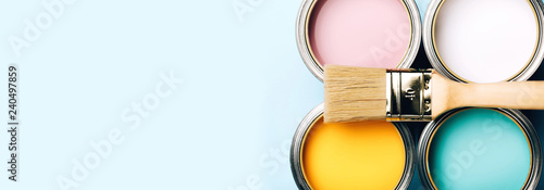 Banner of renovation concept. Brush with wooden handle on opened cans on blue pastel background. Yellow, white, pink, turquoise colors. Macro.