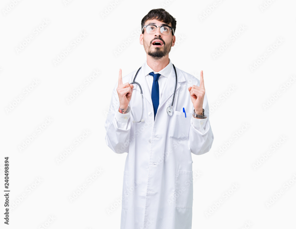 Young doctor man wearing hospital coat over isolated background amazed and surprised looking up and pointing with fingers and raised arms. Young doctor man wearing hospital coat over isolated background amazed and surprised looking up and pointing with fingers and raised arms.