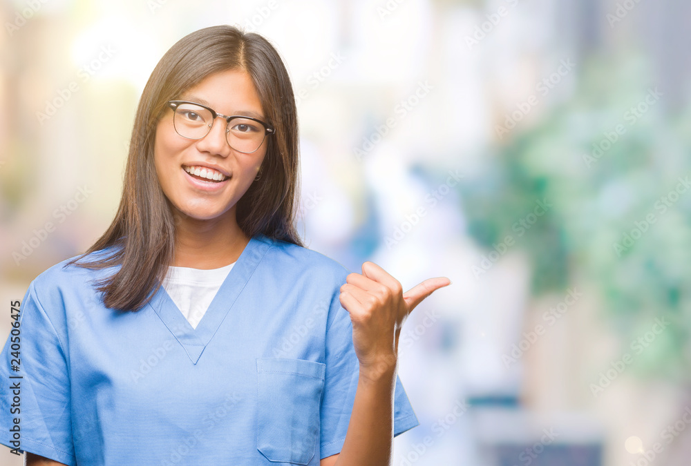 Young asian doctor woman over isolated background smiling with happy face looking and pointing to the side with thumb up.
