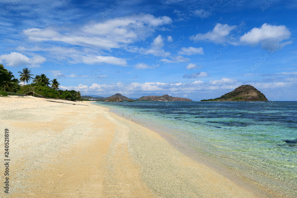 White sand clear beach with palm trees and mountains on cloudy blue sky background at Sumbawa island, Indonesia
