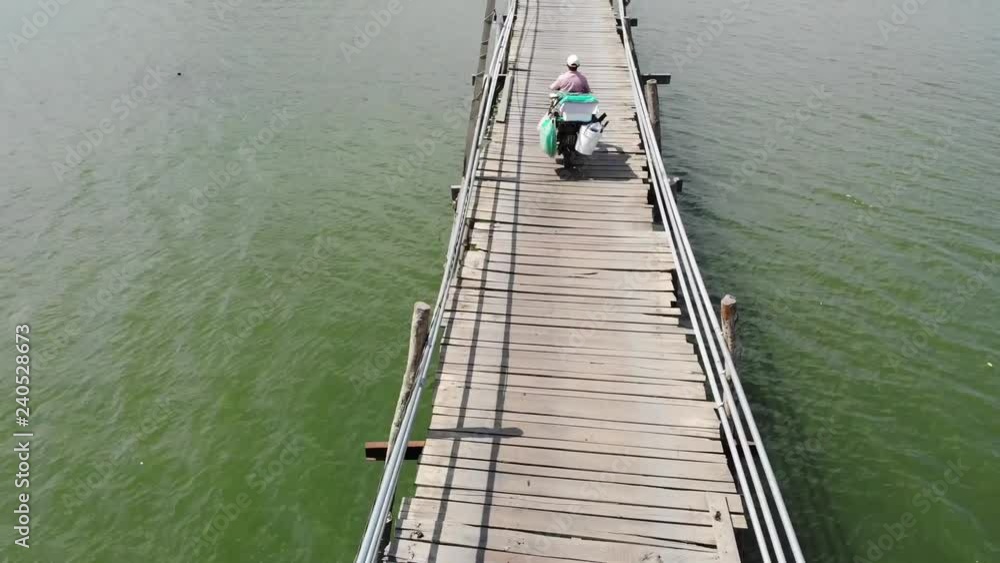 A flying camera follows a motorcyclist riding an old small wooden bridge across a river in an Asian village near Nha Trang Vietnam from a drone. A flying camera follows a motorcyclist riding an old small wooden bridge across a river in an Asian village near Nha Trang Vietnam from a drone.