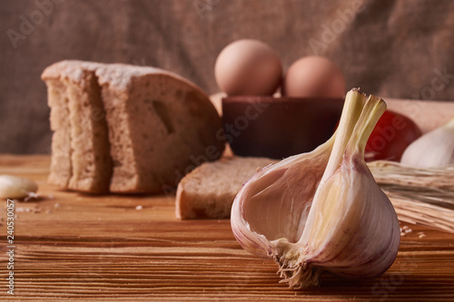 Obraz na plátne garlic on a wooden table with eggs in clay bowl, tomato, battledore and wheat sp