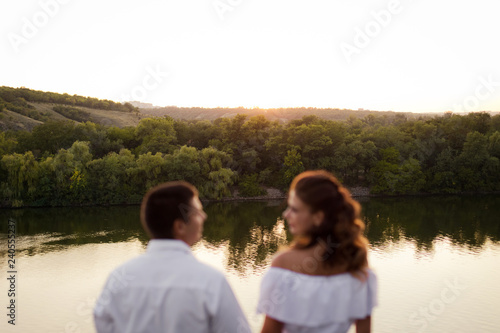 Young beautiful woman in long white dress with her husband on the stones at the river on sunset time