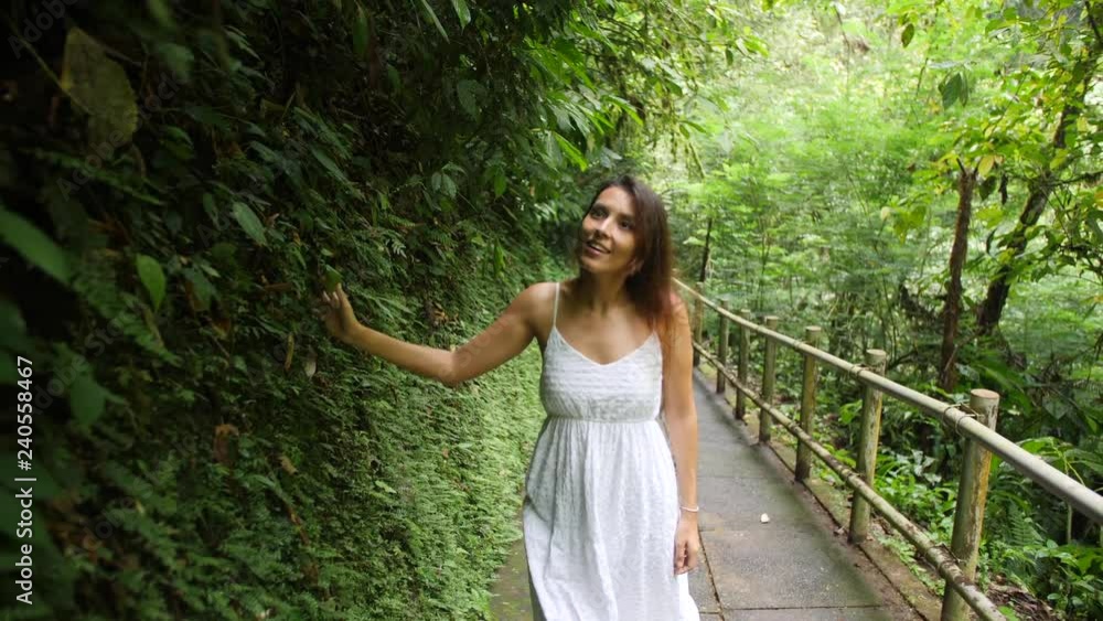Young Happy Mixed Race Girl in White Dress Walking in Jungle Forest, Looking Around and Touching with Hand Tropical Leaves. Calm and Carefree Lifestyle Travel 4K Slowmotion Footage. Bali, Indonesia. Young Happy Mixed Race Girl in White Dress Walking in Jungle Forest, Looking Around and Touching with Hand Tropical Leaves. Calm and Carefree Lifestyle Travel 4K Slowmotion Footage. Bali, Indonesia.