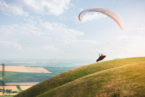 Professional paraglider in a cocoon suit flies high above the ground against the sky and fields