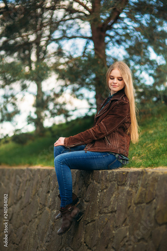 Attrective young girl sits by the embankment. Blond hair and brown jacket. Spring