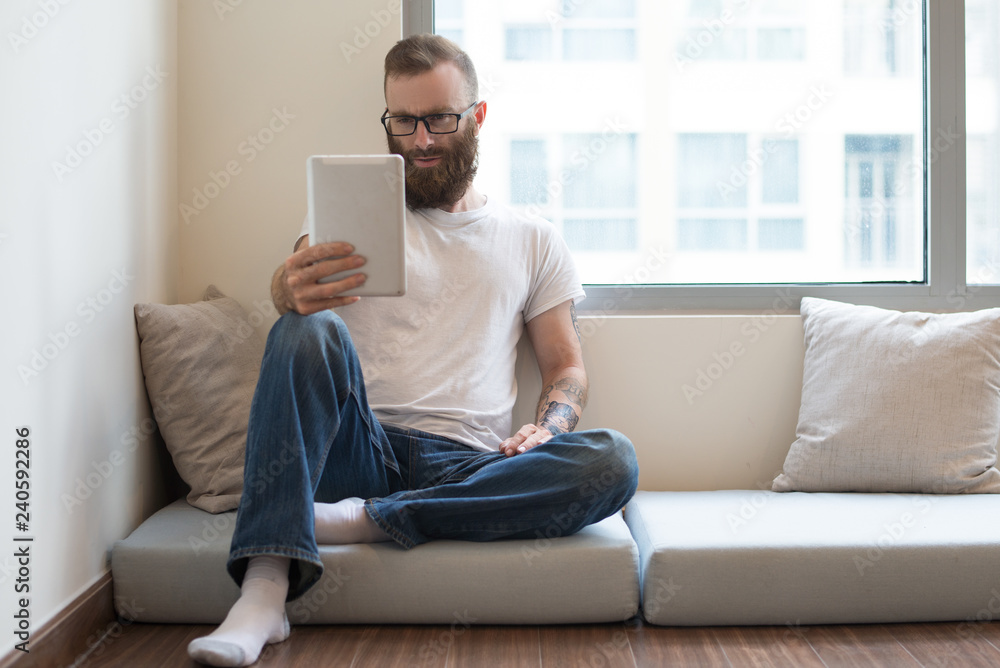 Concentrated bearded man sitting on pillow and using tablet. Serious young guy watching video on gadget. Modern device concept Concentrated bearded man sitting on pillow and using tablet. Serious young guy watching video on gadget. Modern device concept