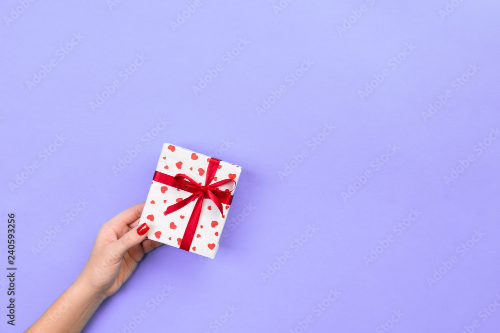 Fototapeta premium Woman hands give wrapped valentine or other holiday handmade present in paper with red ribbon. Present box, red heart decoration of gift on violet table, top view with copy space for you design