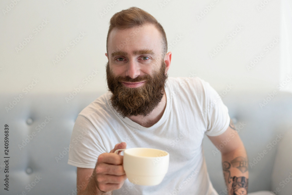 Happy bearded young man in casual enjoying coffee break on couch. Stylish hipster drinking coffee at home. Weekend morning concept Happy bearded young man in casual enjoying coffee break on couch. Stylish hipster drinking coffee at home. Weekend morning concept