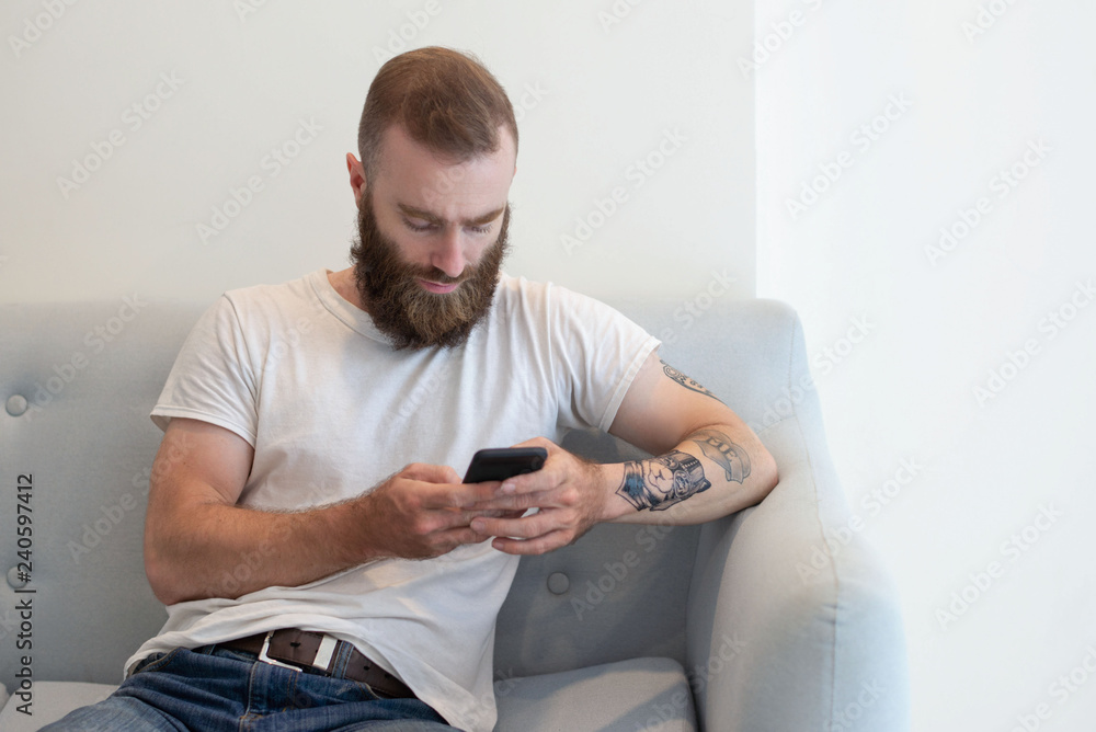 Serious bearded hipster using smartphone in waiting room. Focused young man in casual resting on couch and testing mobile app. Phone using concept