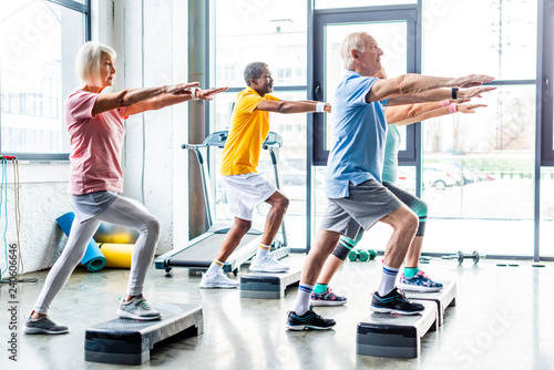 Fototapeta Naklejka Na Ścianę i Meble -  side view of multicultural senior athletes synchronous exercising on step platforms at gym