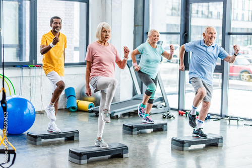 Fototapeta Naklejka Na Ścianę i Meble -  happy multiethnic senior athletes synchronous exercising on step platforms at gym