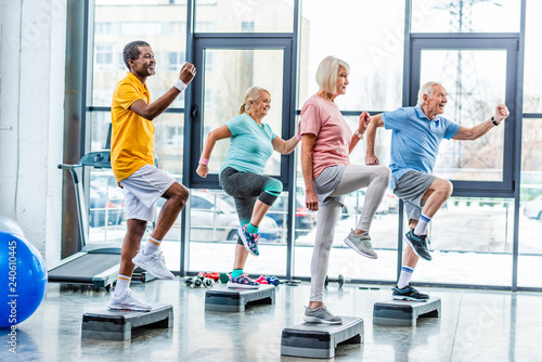 Fototapeta Naklejka Na Ścianę i Meble -  side view of multiethnic senior athletes synchronous exercising on step platforms at gym
