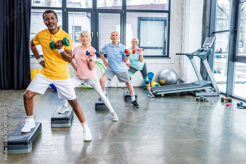 Fototapeta Naklejka Na Ścianę i Meble -  multiethnic senior sportspeople synchronous exercising with dumbbells on step platforms at gym