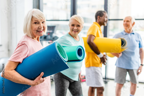 Fototapeta Naklejka Na Ścianę i Meble -  selective focus of senior sportswoman holding fitness mat and her friends standing behind at gym