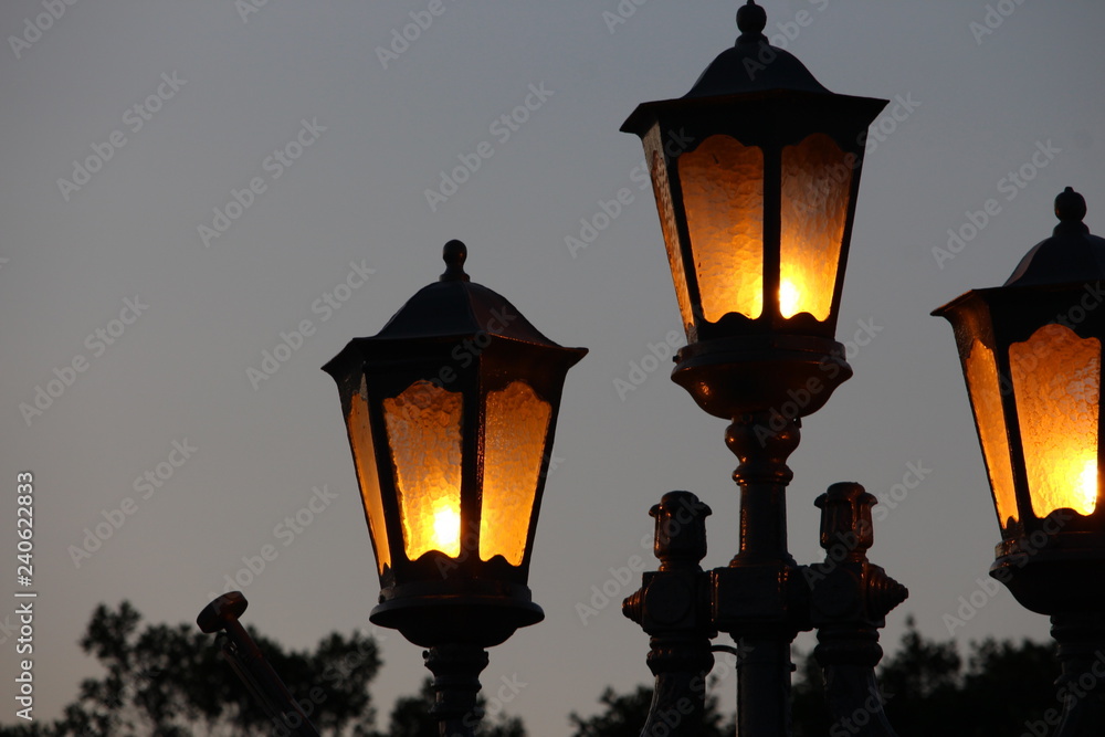 Three lamps, lit, silhouetted against a dusky evening sky, with foliage in the background