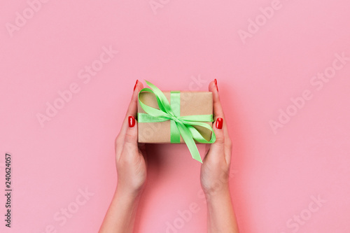 Woman hands give wrapped valentine or other holiday handmade present in paper with green ribbon. Present box, decoration of gift on Coral color table, top view with copy space