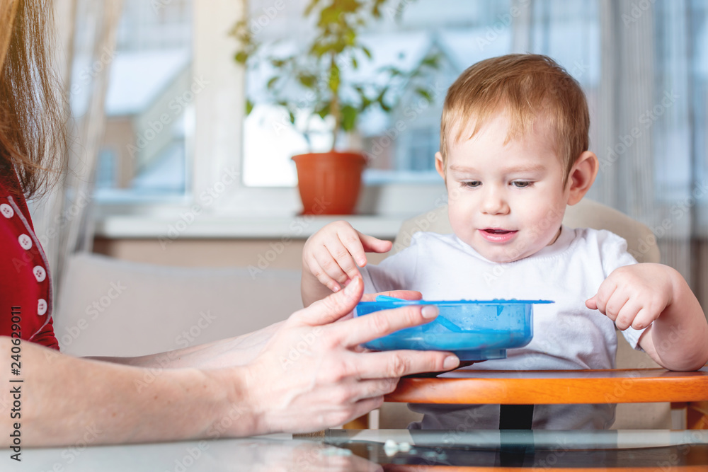 Little baby boy learning to eat with a spoon himself at the kids table in the kitchen. Concept of healthy baby food Little baby boy learning to eat with a spoon himself at the kids table in the kitchen. Concept of healthy baby food