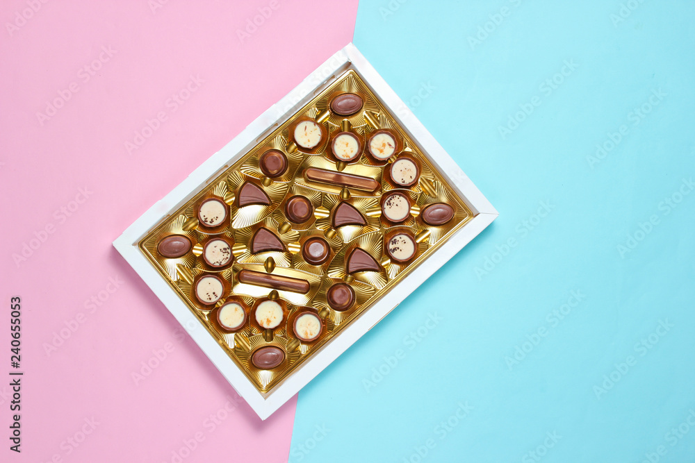 Open box with golden tray of variety of chocolate candies on plue pink pastel background. Top view, minimalism. Open box with golden tray of variety of chocolate candies on plue pink pastel background. Top view, minimalism.
