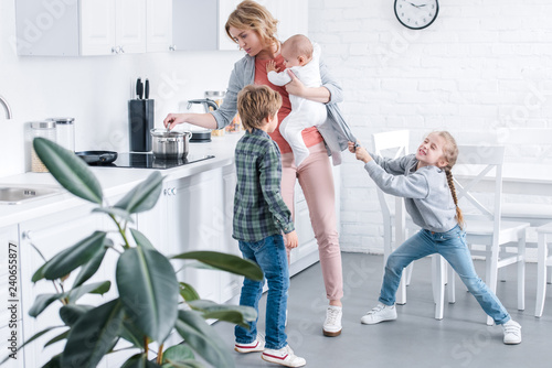 tired mother holding infant child and cooking while naughty children playing in kitchen photo