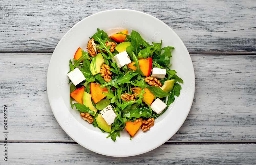 Arugula salad, persimmon, walnuts, avocados, feta cheese on a white plate on a wooden table Arugula salad, persimmon, walnuts, avocados, feta cheese on a white plate on a wooden table
