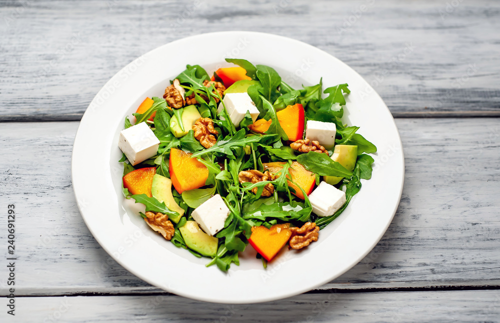 Arugula salad, persimmon, walnuts, avocados, feta cheese on a white plate on a wooden table