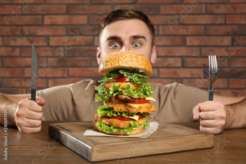 Fototapeta Young hungry man with cutlery eating huge burger at table