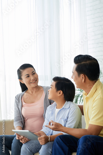 Cheerful Vietnamese family sitting on sofa and discussing new application on tablet computer