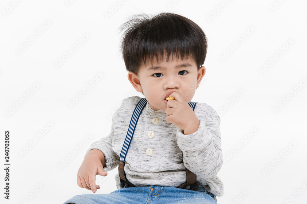 Studio portrait of cute, adorable, Asian toddler boy wearing denim overalls, long sleeve T-shirt, orange shoes, sitting spread legs on the floor, enjoy eating snack, on isolated white background Studio portrait of cute, adorable, Asian toddler boy wearing denim overalls, long sleeve T-shirt, orange shoes, sitting spread legs on the floor, enjoy eating snack, on isolated white background
