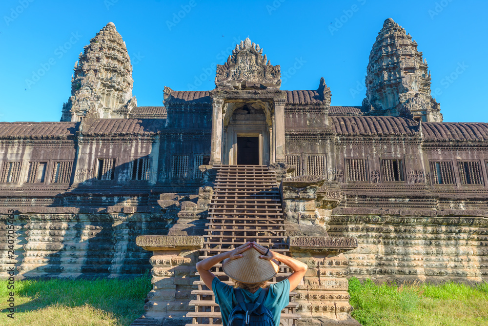 Fototapeta premium One tourist visiting Angkor Wat ruins at sunrise, travel destination Cambodia. Woman with traditional hat and raised arms, rear view, main facade staircase gate and towers.