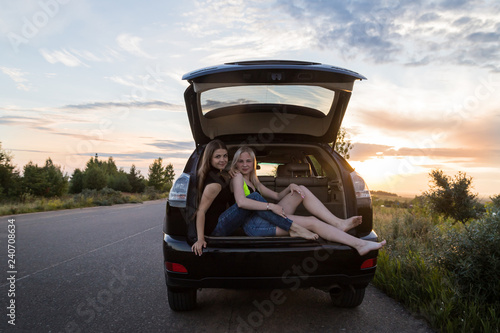 Two girls sitting in the trunk of a car. Friends relax in the summer evening and sunset behind them © keleny