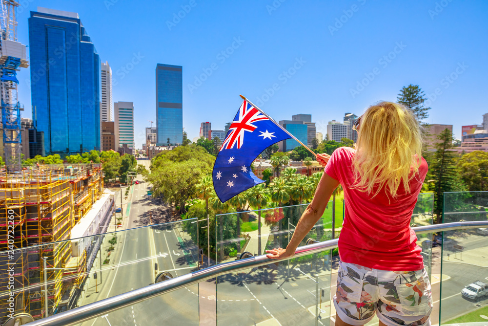Blonde woman with Australian flag on the Bell Tower observation deck in Perth, Western Australia. Blue sky in a summer season. Elizabeth Quay activity. Caucasian tourist enjoys city views.
