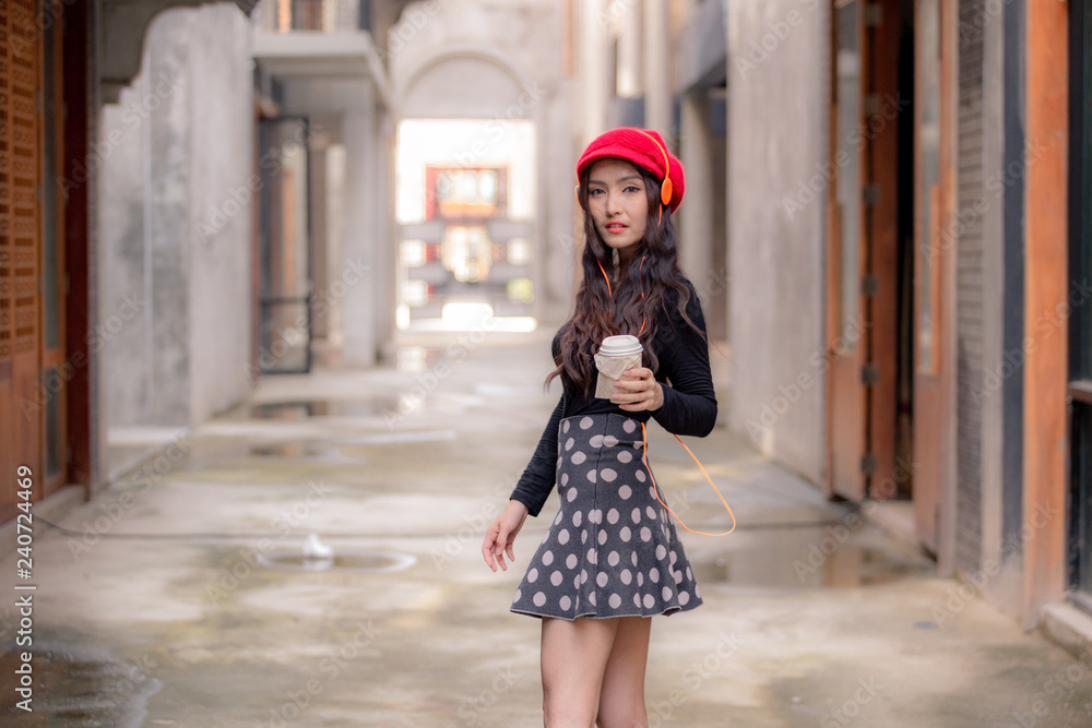 A beautiful charming asian girl turn back herself while enjoy listening music with headphone and holding a cup of coffee Feeling look happy and relax on walking street. Portrait lady winter concept.