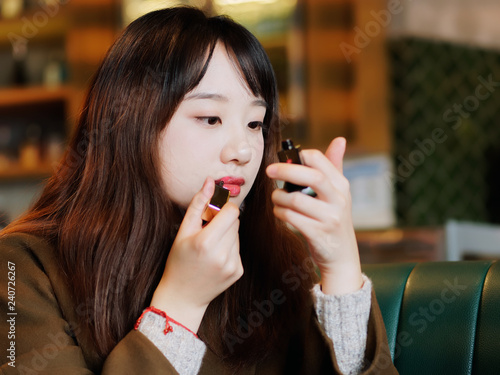 Portrait of charming Chinese young woman applying lipstick looking at small mirror hold in hand. Beautiful girl makes makeup and get ready before date.
