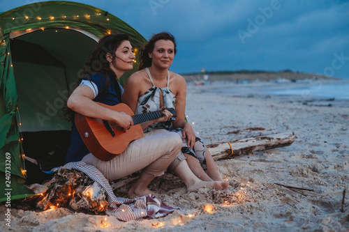 Young loving couple while camping on the seashore a summer evening at sunset. One woman plays the guitar while the other is listening to her