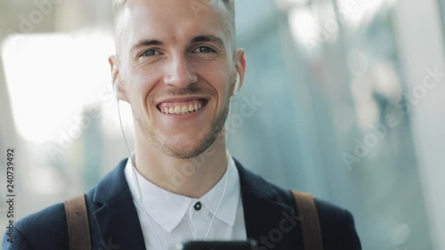 Young attractive businessman standing near business center with smartphone, coffee and headphones. He looking into the camera photo