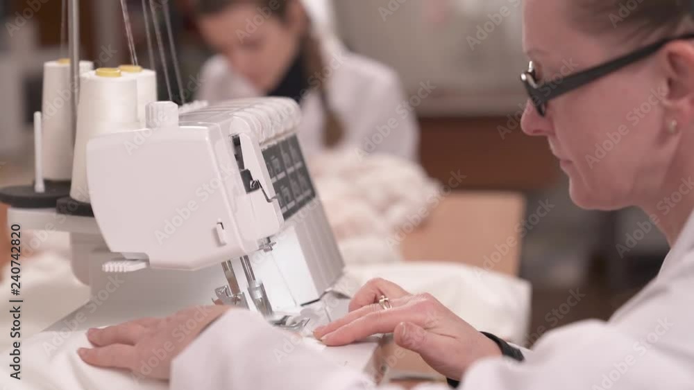 A close up side view of a seamstress finishing stitching white fabric on the sewing machine. Another tailor is sitting at the table and sewing beads down the veiling cloth to make a wedding dress A close up side view of a seamstress finishing stitching white fabric on the sewing machine. Another tailor is sitting at the table and sewing beads down the veiling cloth to make a wedding dress