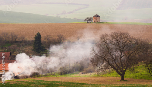 Rural Pastoral Spring Landscape With Old Windmill Without Blades And Plowed Land On Background Of The Village And Thick White Smoke.European Ancient Windmill Or Defense Tower On The Brown Wavy Field