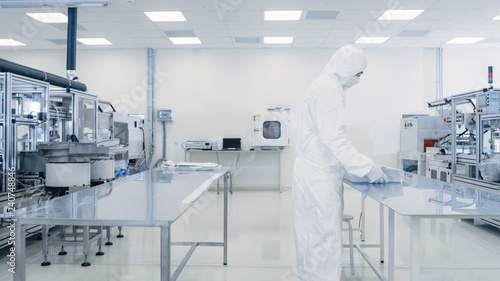 Scientist in Protective Suit Working with Case of Finished Product Through Laboratory. Workers in the Facility with Modern Industrial Machinery. Product Manufacturing Process: Biotechnology.